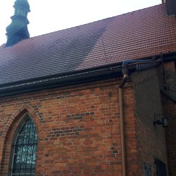 Renodach Serwis - Fragment kościoła z czerwonej cegły z widocznym dachem, rynną i wieżą z krzyżem; część dachu jest wyraźnie czystsza od reszty.