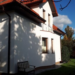 Renodach Serwis - Boczny widok domu z jasną elewacją, brązowymi rynnami i czerwonym dachem, z widocznymi cieniami drzew na ścianie i krzesłem ogrodowym w pobliżu.