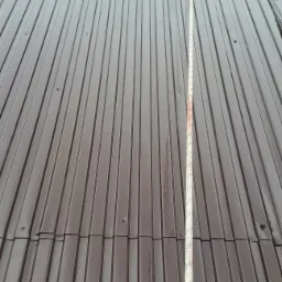 Ciemnobrązowy dach z blachy trapezowej, widoczna pionowa smuga brudu po lewej stronie liny zabezpieczającej, sugerująca niedawne czyszczenie części dachu.