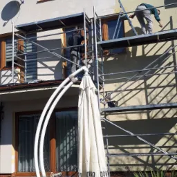 Dwóch pracowników na rusztowaniu maluje elewację domu jednorodzinnego, widoczne fragmenty pomalowanej ściany, w tle błękitne niebo, w pierwszym planie złożony parasol ogrodowy.