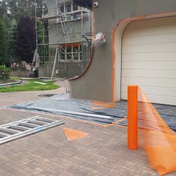 Renodach Serwis - Elewacja domu w trakcie prac: szare ściany, rusztowania, zabezpieczenia pomarańczową siatką. Widoczny fragment ogrodu i kostka brukowa.