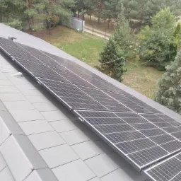 Panel słoneczny na szarym dachu z blachy trapezowej, widok z góry na zielony ogród z drzewami iglastymi w tle.