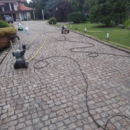 Czyszczenie kostki brukowej na podjeździe do domu jednorodzinnego z wykorzystaniem myjek ciśnieniowych; widoczne urządzenia, węże i mokra powierzchnia kostki.