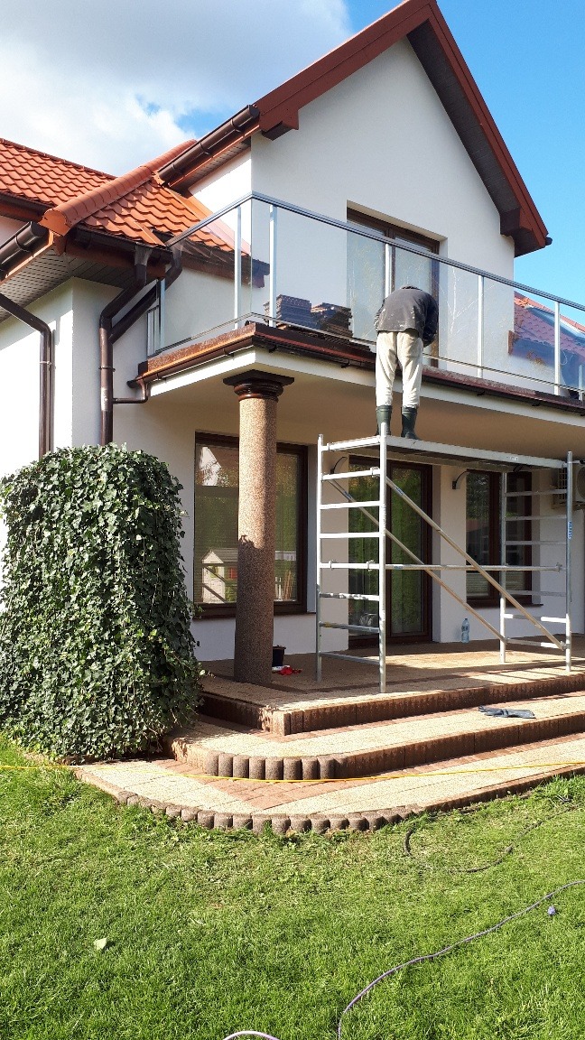 Pracownik na rusztowaniu przy balkonie domu jednorodzinnego z brązowym dachem; widoczne przygotowane płytki na balkonie z szklaną balustradą.
