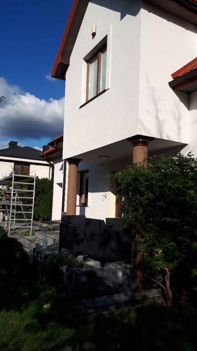 Jasna elewacja domu jednorodzinnego z kolumnami w odcieniach brązu podtrzymującymi zadaszenie tarasu, widoczne rusztowanie i meble ogrodowe.