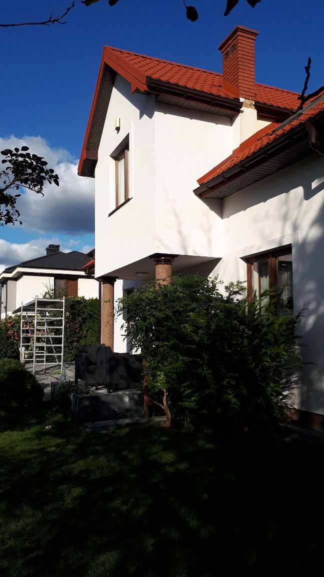 Biała elewacja domu z czerwoną dachówką i kominem w słoneczny dzień, widoczne kolumny podtrzymujące zadaszenie tarasu i drabina oparta o ścianę drugiego budynku w tle.