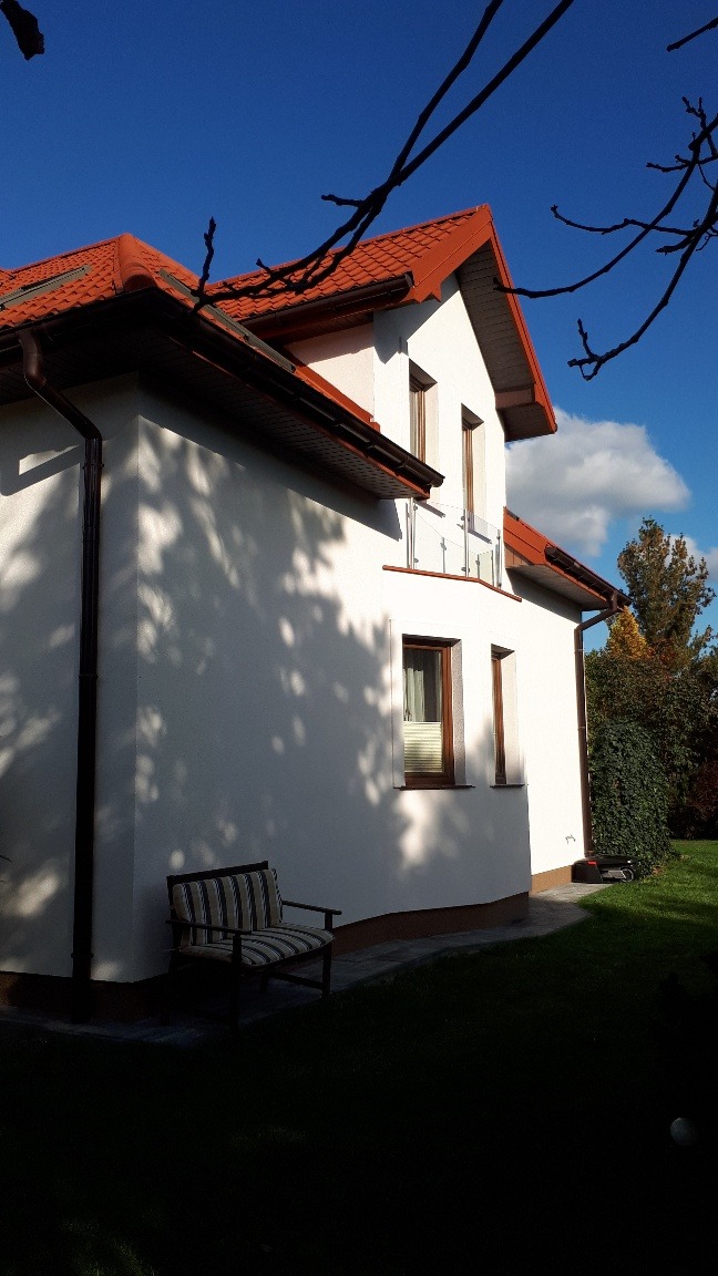 Boczny widok domu z jasną elewacją, brązowymi rynnami i czerwonym dachem, z widocznymi cieniami drzew na ścianie i krzesłem ogrodowym w pobliżu.