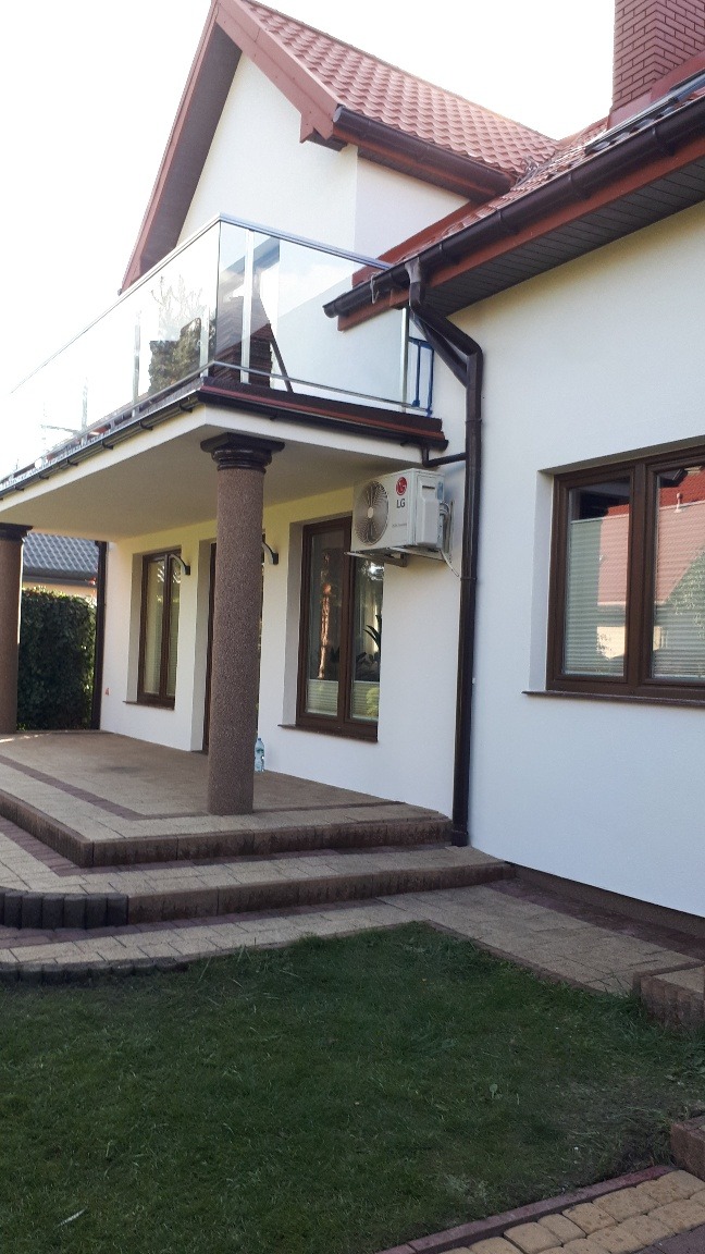 Jasna elewacja domu jednorodzinnego z brązowymi oknami, rynnami i kolumnami werandy, widoczny klimatyzator LG na ścianie.