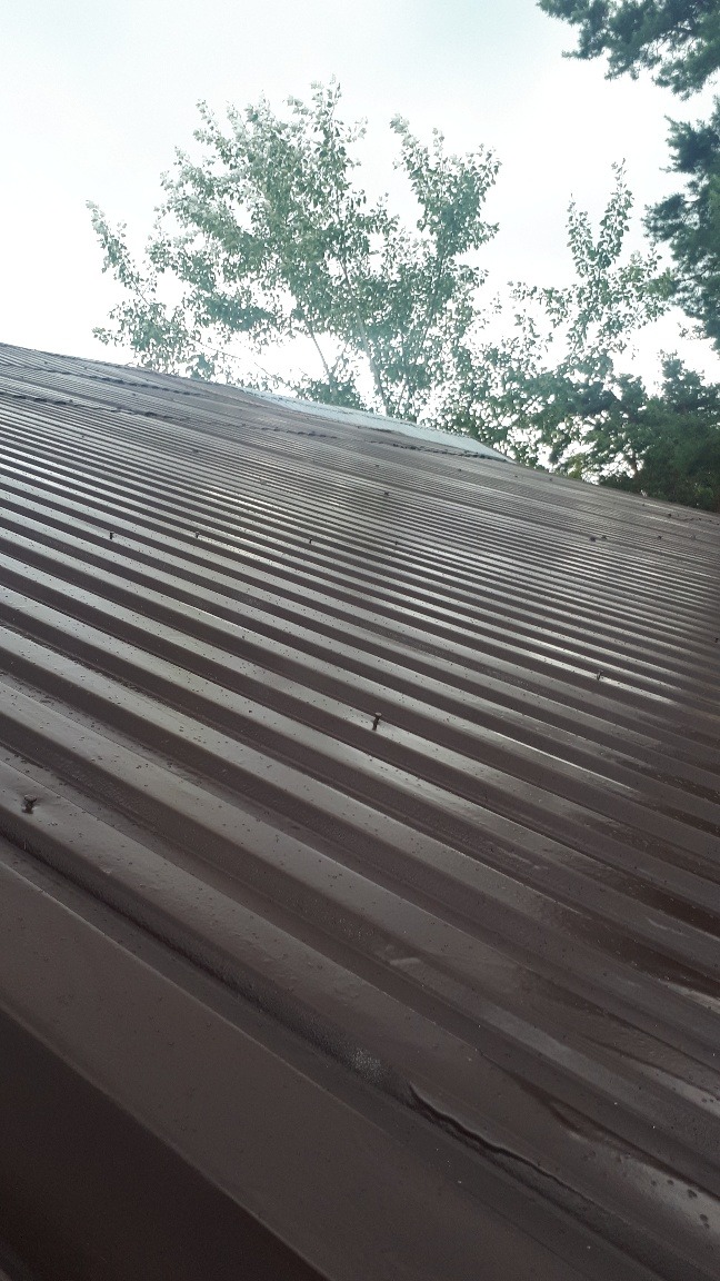 Metalowy dach w kolorze ciemnego brązu, z widocznymi śladami po niedawnym malowaniu, w tle drzewa i pochmurne niebo.