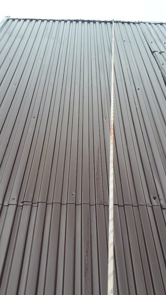 Ciemnobrązowy dach z blachy trapezowej, widoczna pionowa smuga brudu po lewej stronie liny zabezpieczającej, sugerująca niedawne czyszczenie części dachu.