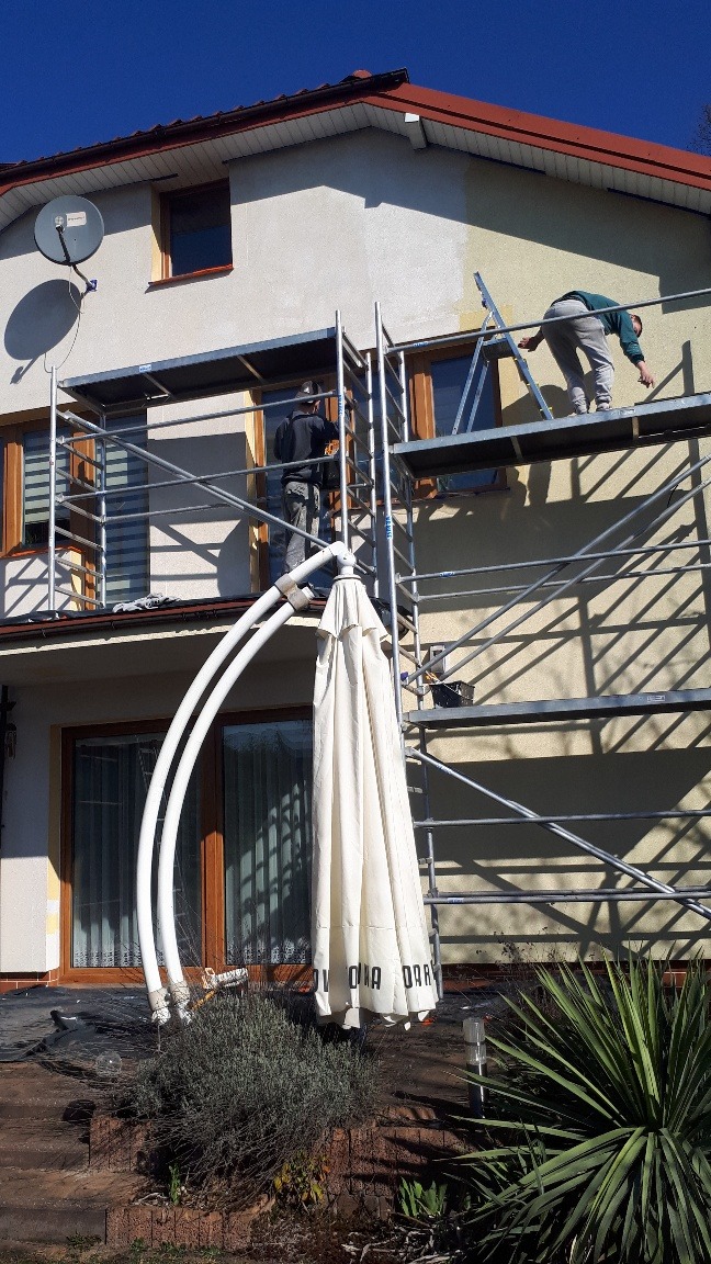 Dwóch pracowników na rusztowaniu maluje elewację domu jednorodzinnego, widoczne fragmenty pomalowanej ściany, w tle błękitne niebo, w pierwszym planie złożony parasol ogrodowy.