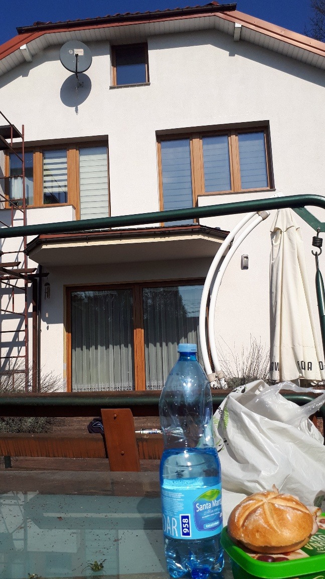 Dom z białą elewacją, rusztowaniem i anteną satelitarną, częściowo zasłonięty przez butelkę wody, bułkę i torbę na pierwszym planie.