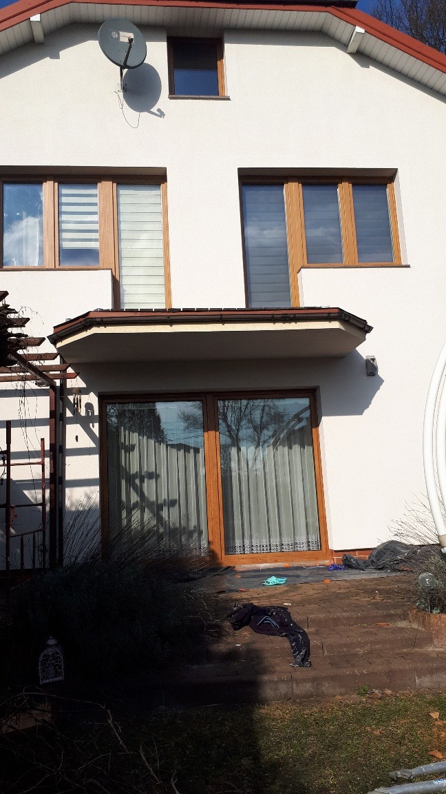 Dom jednorodzinny w Płońsku z jasną elewacją i drewnianą stolarką okienną. Widoczne ślady prac na schodach prowadzących do wejścia, sugerujące niedawne prace na elewacji.