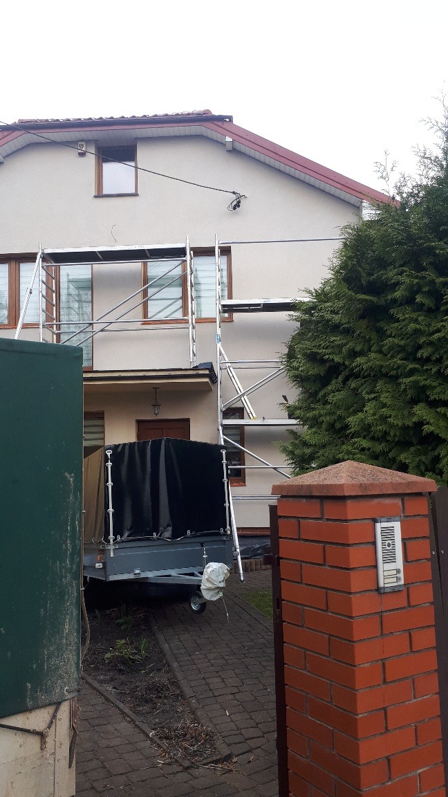 Elewacja domu z ustawionym rusztowaniem, widoczny fragment przyczepki z plandeką i ceglany słupek z domofonem.