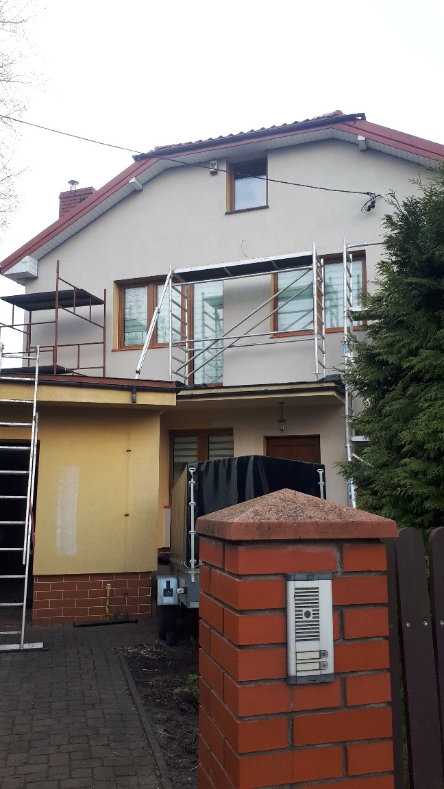 Dom jednorodzinny w trakcie malowania elewacji, widoczne rusztowania i fragment przyczepki z plandeką.