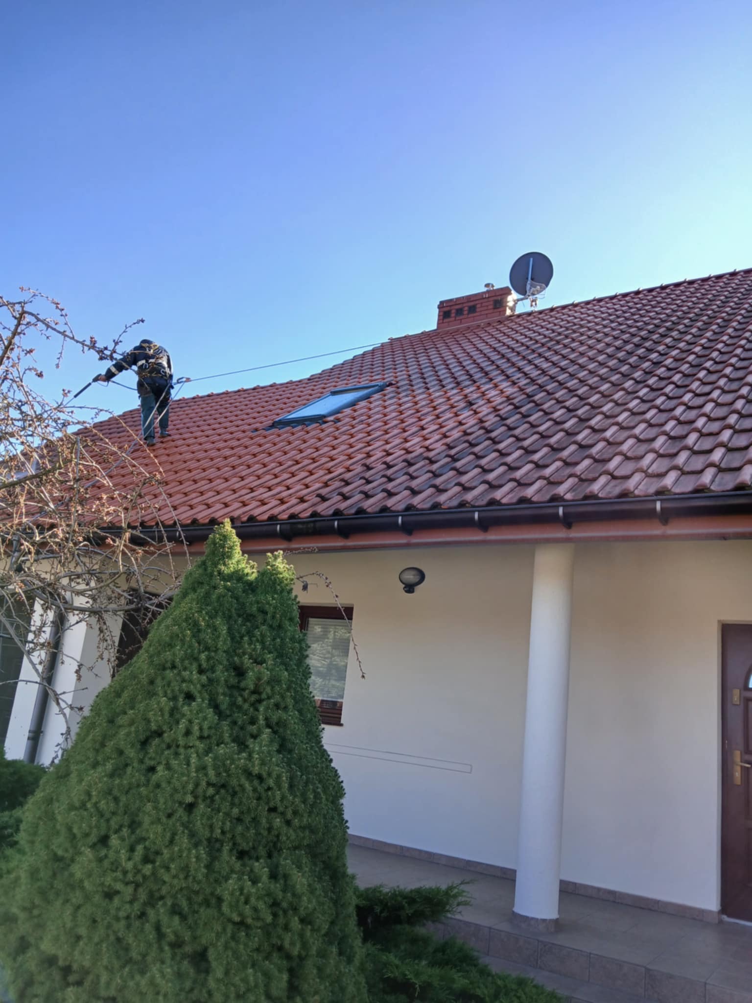 Pracownik zabezpieczony linami na dachu z czerwonej dachówki, z anteną satelitarną w tle, trzyma lancę do czyszczenia, widok z dołu na dom z kolumnami i zielonym iglakiem.