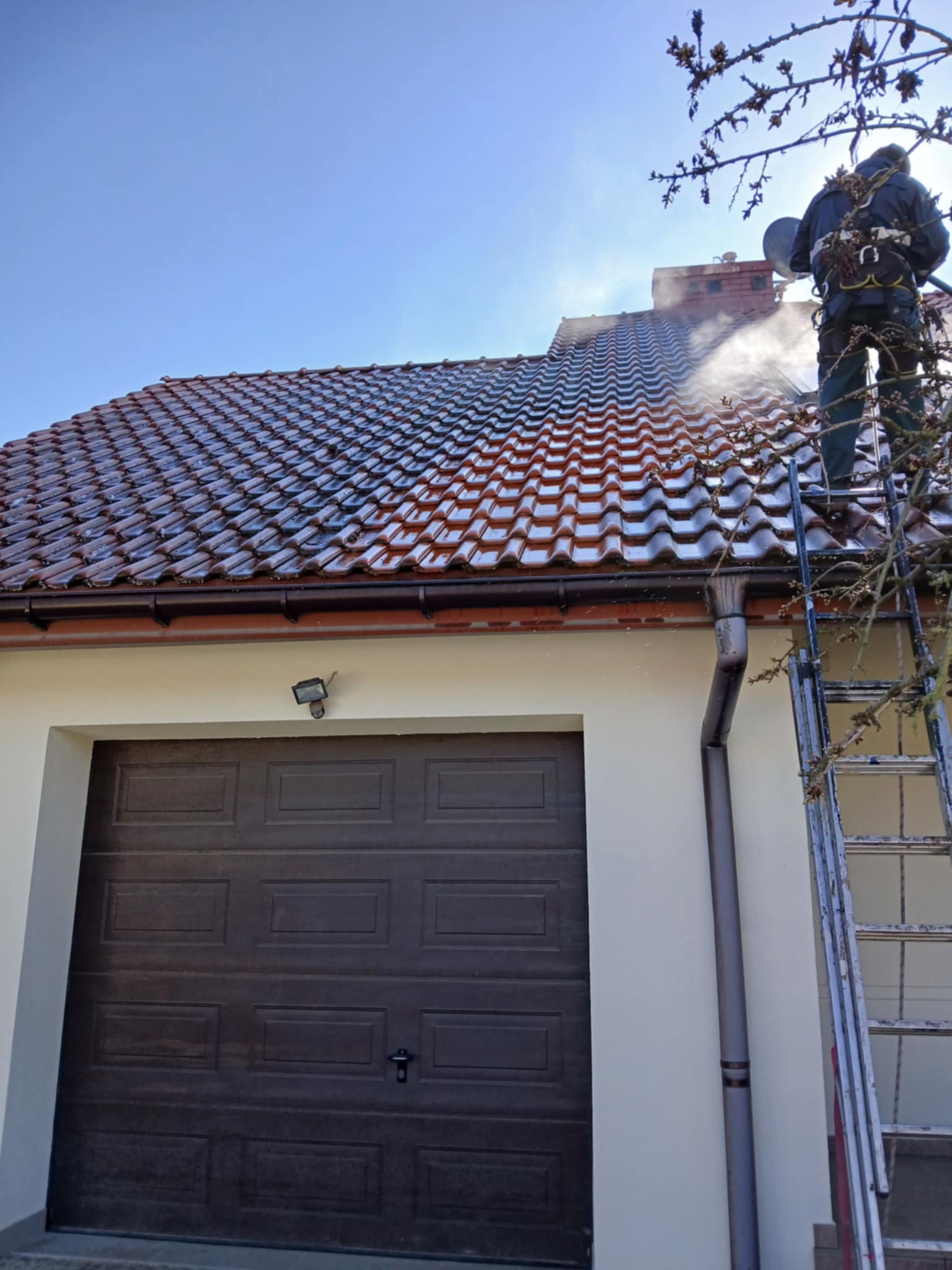 Pracownik w uprzęży na drabinie czyści dach z cegły ceramicznej za pomocą myjki ciśnieniowej; widoczny budynek z brązową bramą garażową i rynnami.