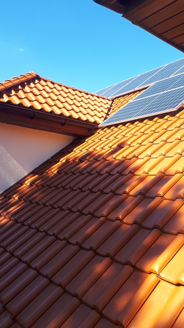 Czerwona dachówka ceramiczna na dachu domu z zamontowanymi panelami słonecznymi, widok z dołu pod kątem, oświetlona promieniami słonecznymi.