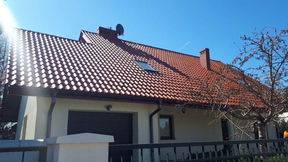 Dom jednorodzinny z czerwoną dachówką, widoczny komin, okno dachowe i antena satelitarna, boczna elewacja w jasnym kolorze, rynna odprowadzająca wodę.