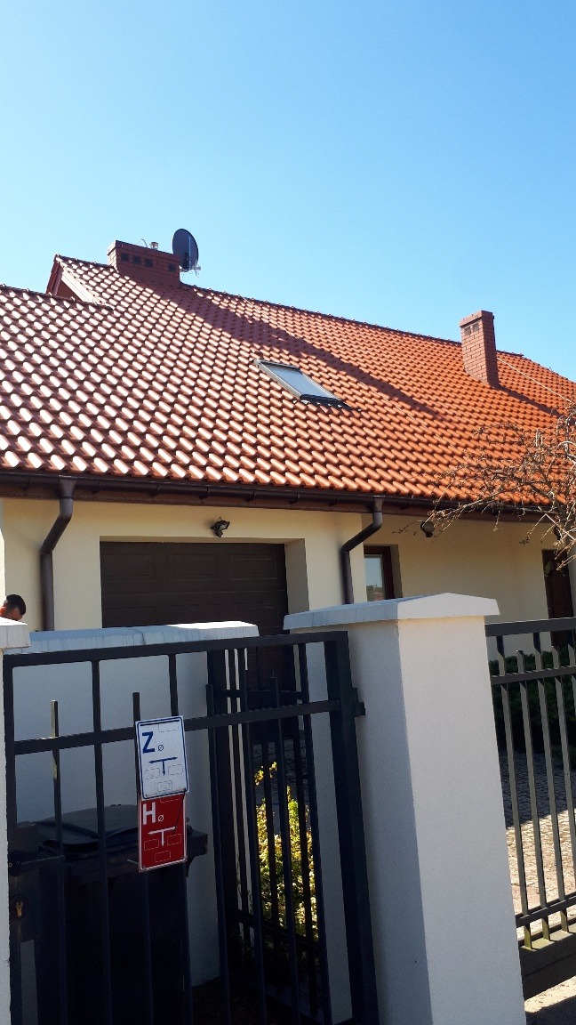 Dom jednorodzinny z czerwoną dachówką, widoczne rynny, okno dachowe, komin z anteną satelitarną, ogrodzenie z bramą i słupkami, oraz tabliczki na ogrodzeniu.