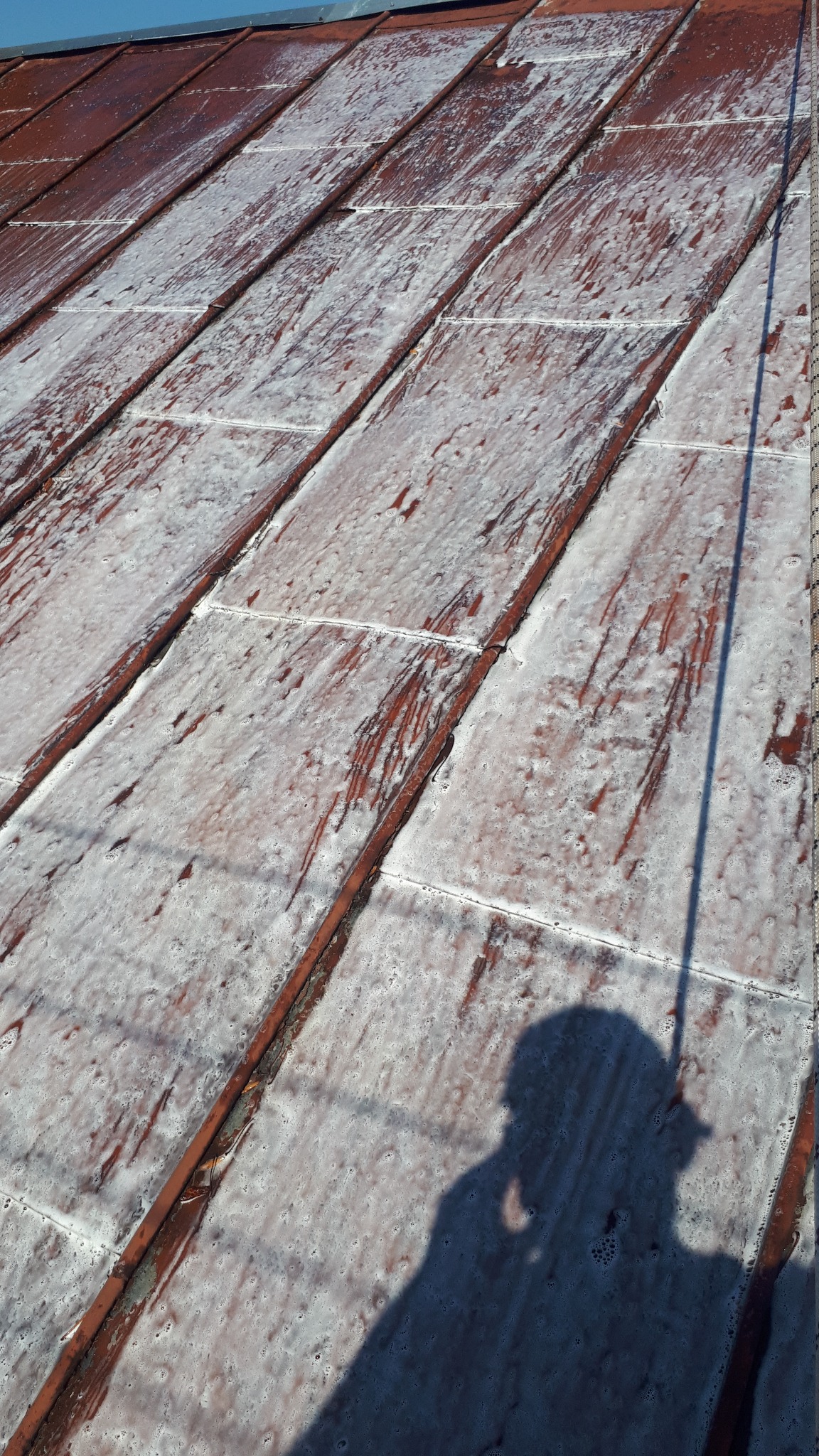 Czerwony dach pokryty pianą czyszczącą, cień osoby z lancą do mycia. Widoczne ślady spływającej piany. Dachówka trapezowa.