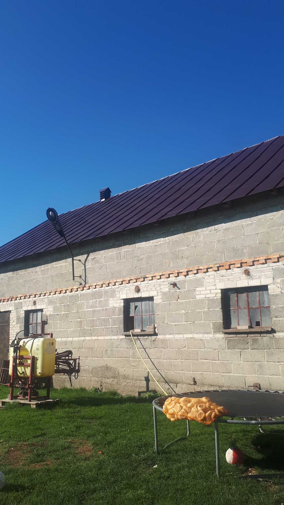 Budynek z szarego pustaka z brązowym dachem z blachy, żółty opryskiwacz i wąż przy ścianie. Na trampolinie leżą siatki z cebulą. Błękitne niebo.
