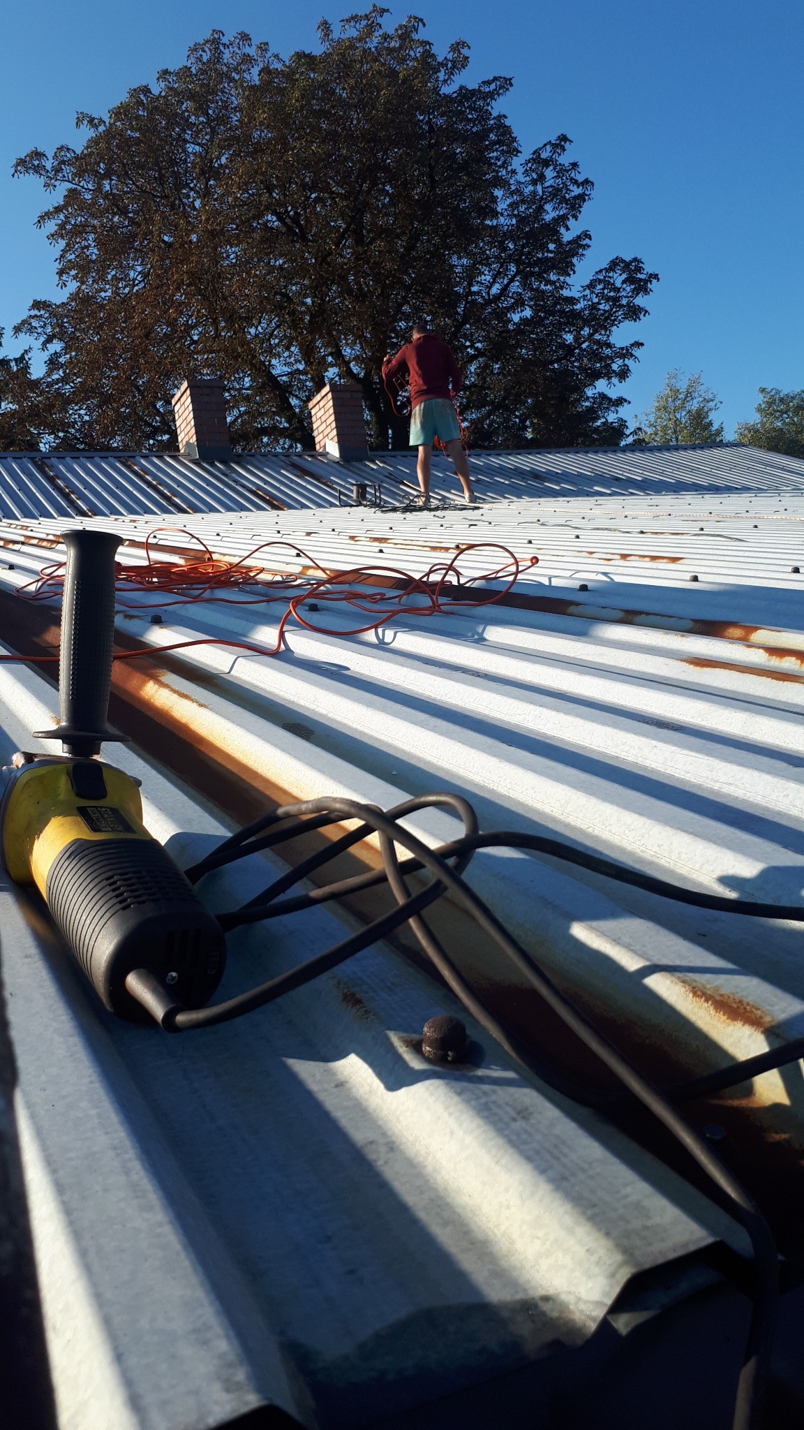 Praca na dachu z blachy falistej: mężczyzna w oddali, narzędzia (szlifierka kątowa) i kable na pierwszym planie, drzewo i kominy w tle, oświetlenie słoneczne.