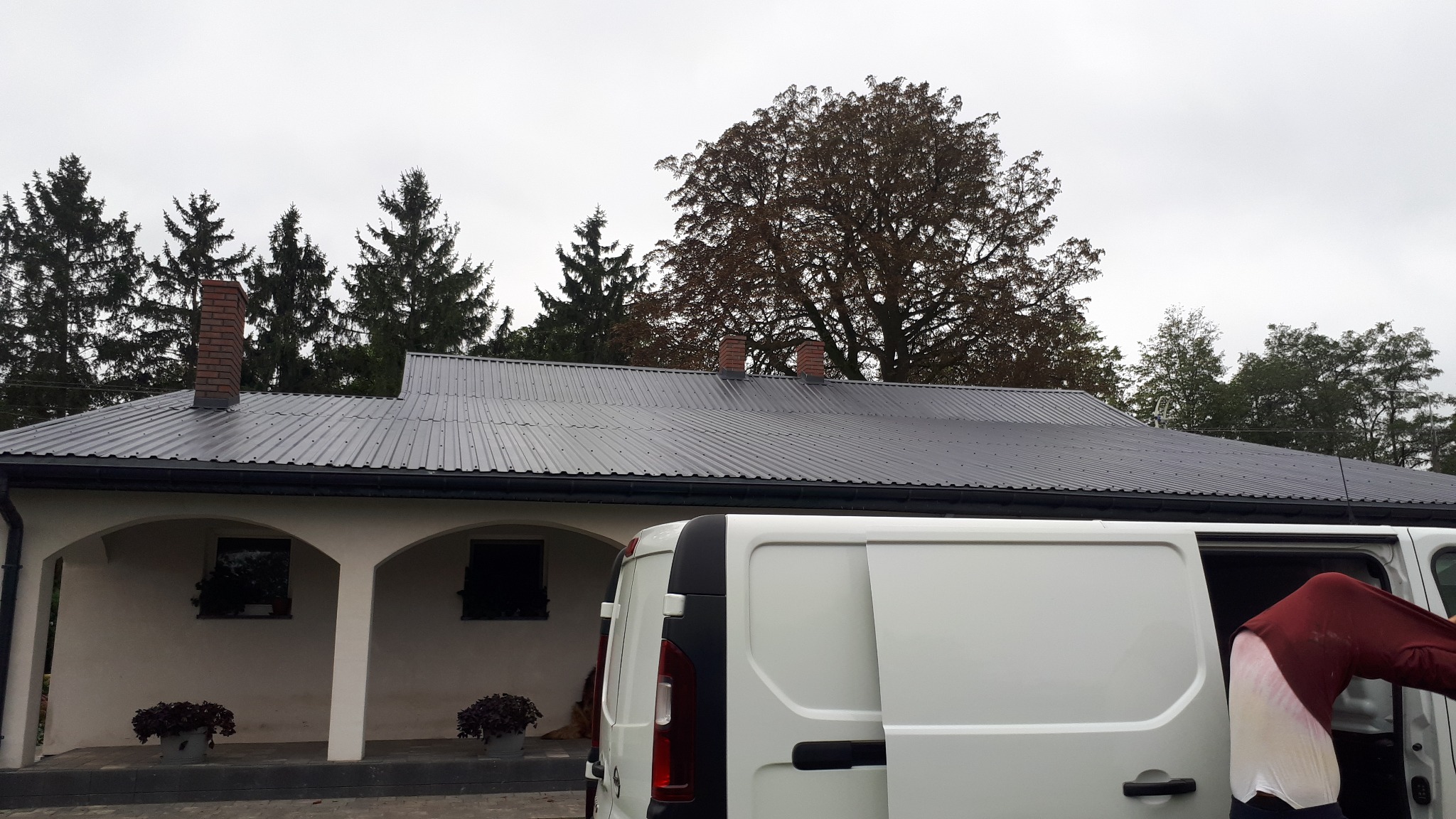 Dom z ciemnoszarym, metalowym dachem po renowacji, widoczny zza białego samochodu dostawczego. W tle drzewa i pochmurne niebo.