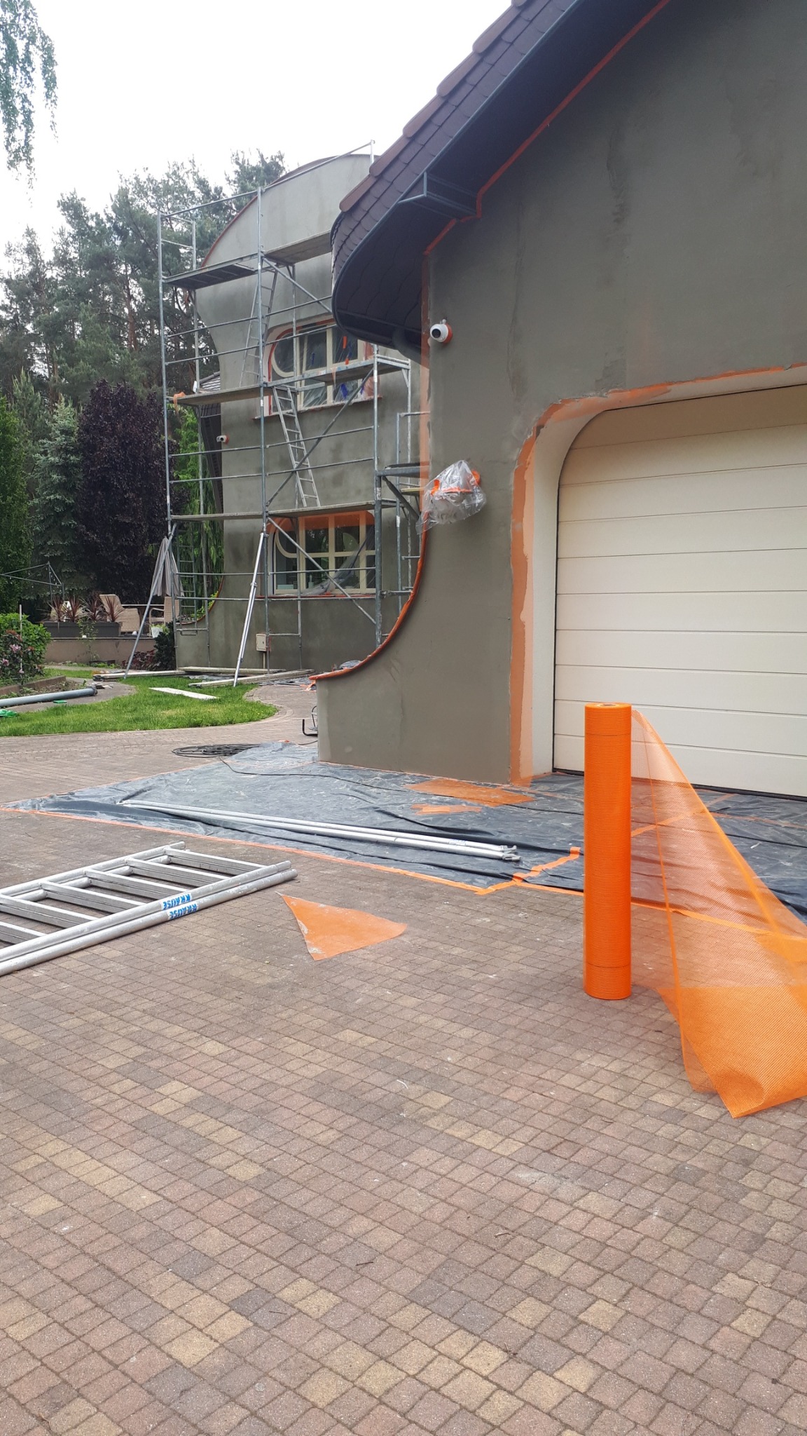 Elewacja domu w trakcie prac: szare ściany, rusztowania, zabezpieczenia pomarańczową siatką. Widoczny fragment ogrodu i kostka brukowa.