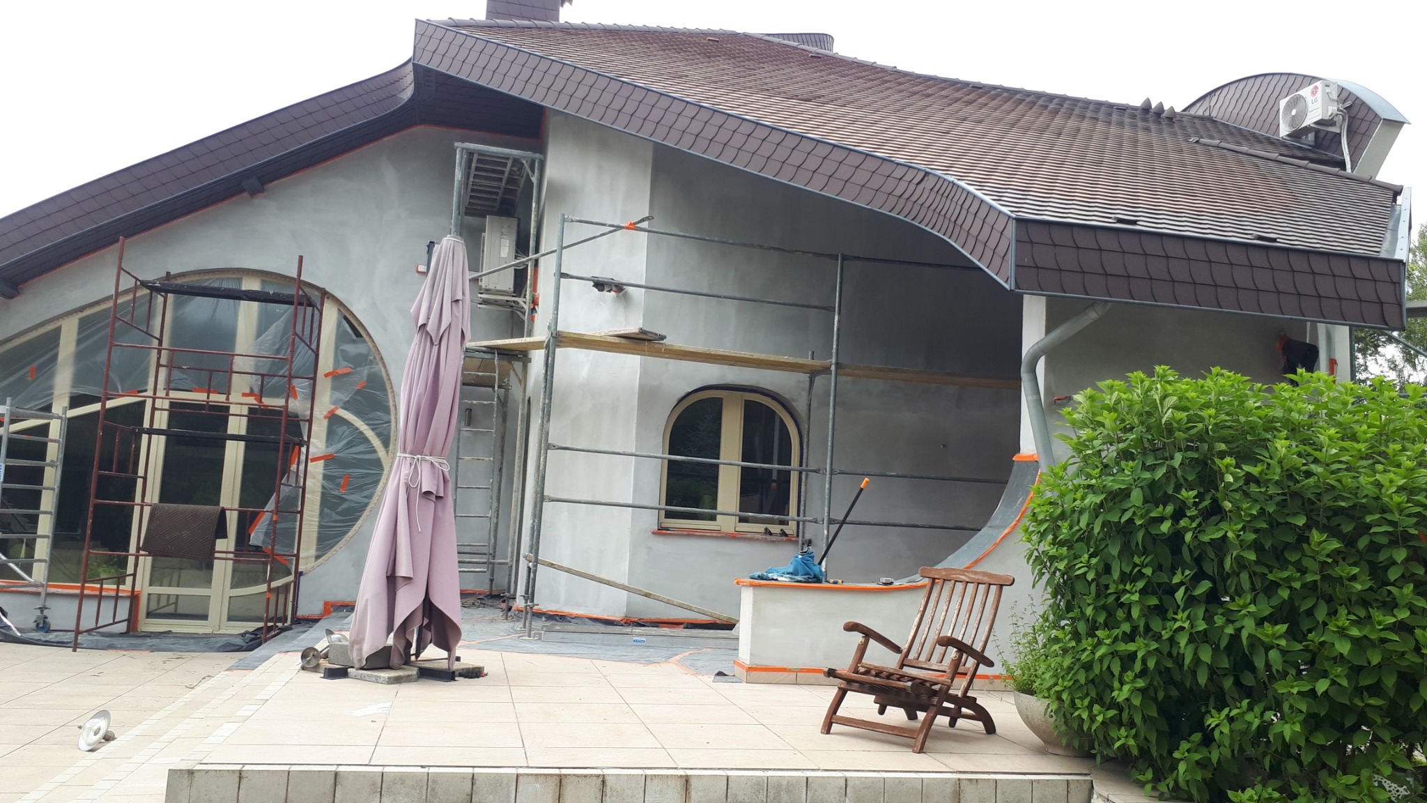 Dom w trakcie malowania elewacji, widoczne rusztowania, zabezpieczone okna folią, przygotowane narzędzia i drewniane krzesła na tarasie.