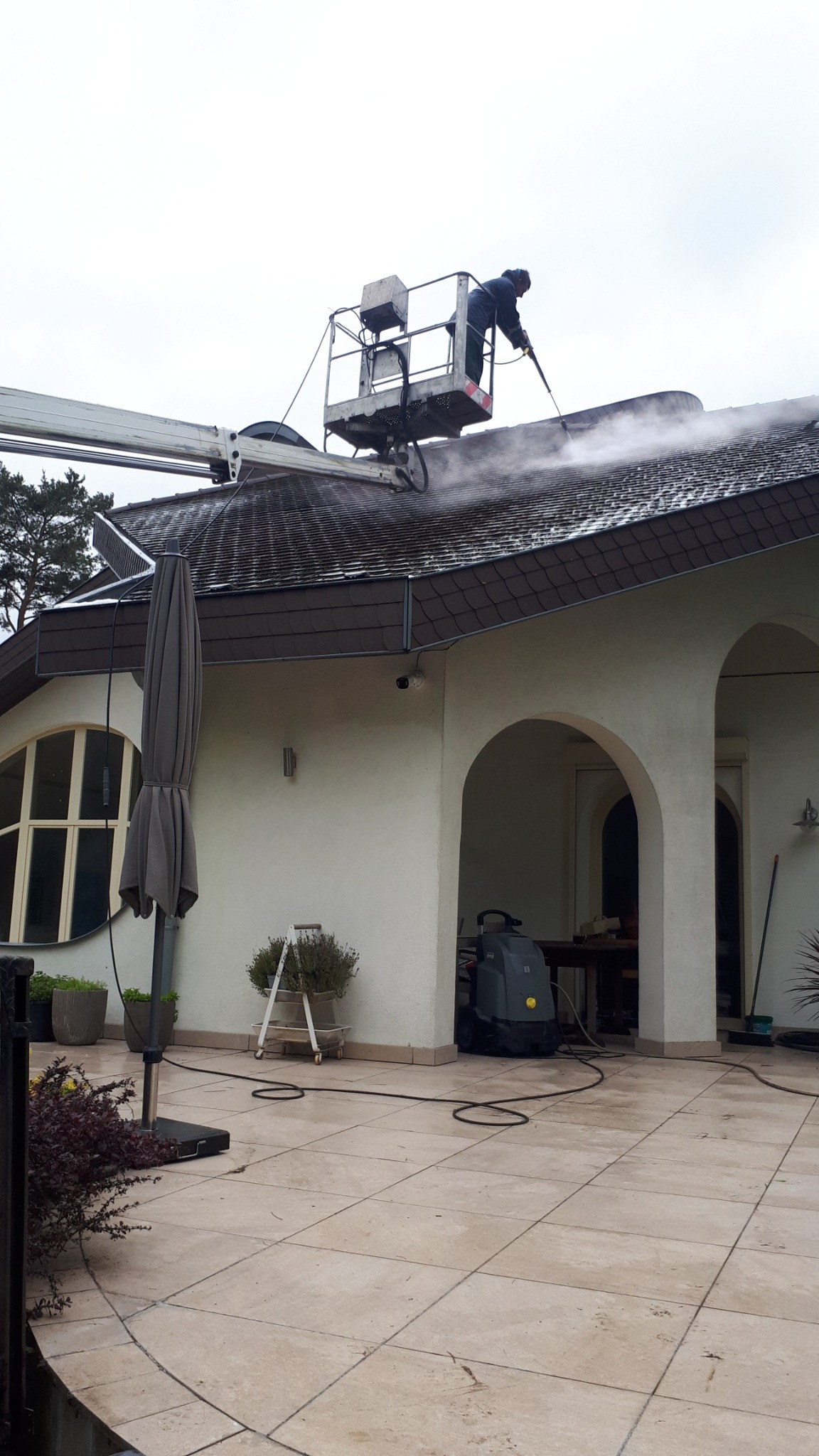 Pracownik na podnośniku czyści dach domu z brązową dachówką za pomocą myjki ciśnieniowej. Widoczny budynek z białymi ścianami i łukowymi oknami.