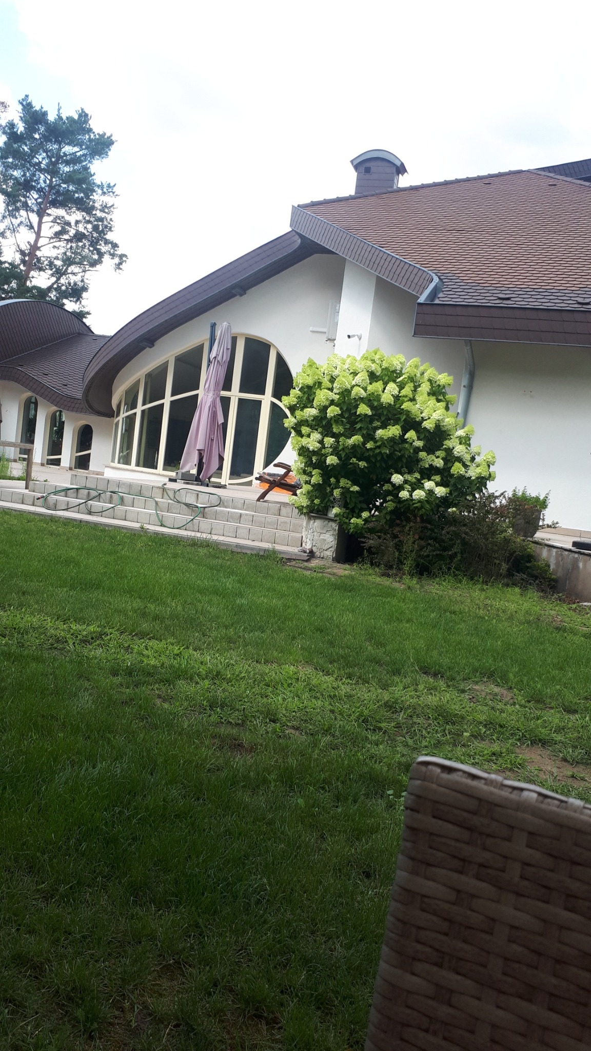 Biała elewacja domu z brązowym dachem, widoczna z poziomu trawnika. Duże okna łukowe, schody wejściowe i krzew z białymi kwiatami. Częściowo widoczny fotel z rattanu.