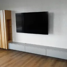 Nowoczesny salon z drewnianą podłogą, szafką RTV w kolorze szarym, wiszącym telewizorem i wysoką, drewnianą szafką z pionowymi panelami i przeszklonymi półkami.