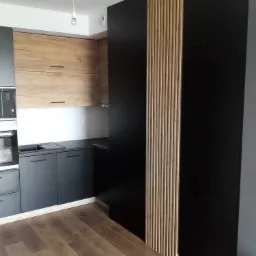 Nowoczesna kuchnia z czarnymi szafkami i drewnianymi akcentami, widok na zabudowę kuchenną z drewnianymi lamelami dekoracyjnymi i sprzętem AGD w zabudowie.