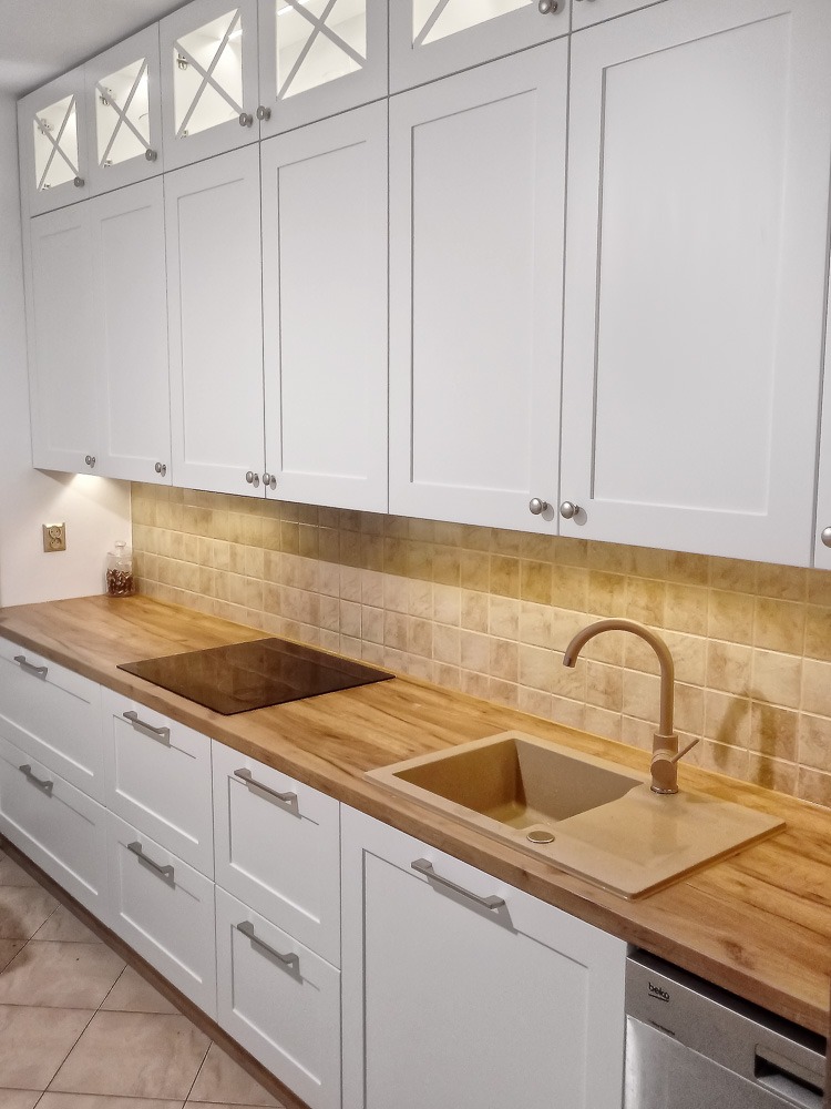 Jasna kuchnia z białymi szafkami z frezowanymi frontami i drewnianym blatem, beżowy zlew i bateria, górne szafki z przeszklonymi frontami z krzyżowym podziałem.