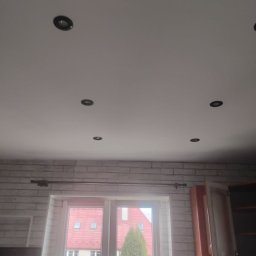 Rafał Niewęgłowski - Biały sufit z sześcioma punktowymi lampami, ściana z białej cegły, okno z widokiem na budynek z czerwonym dachem i zielone drzewa. Częściowo widoczna szafka.