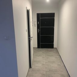 Rafał Niewęgłowski - Korytarz po remoncie: szare płytki podłogowe, białe ściany, czarne drzwi wejściowe z poziomymi srebrnymi paskami, otwarte białe drzwi po lewej stronie.