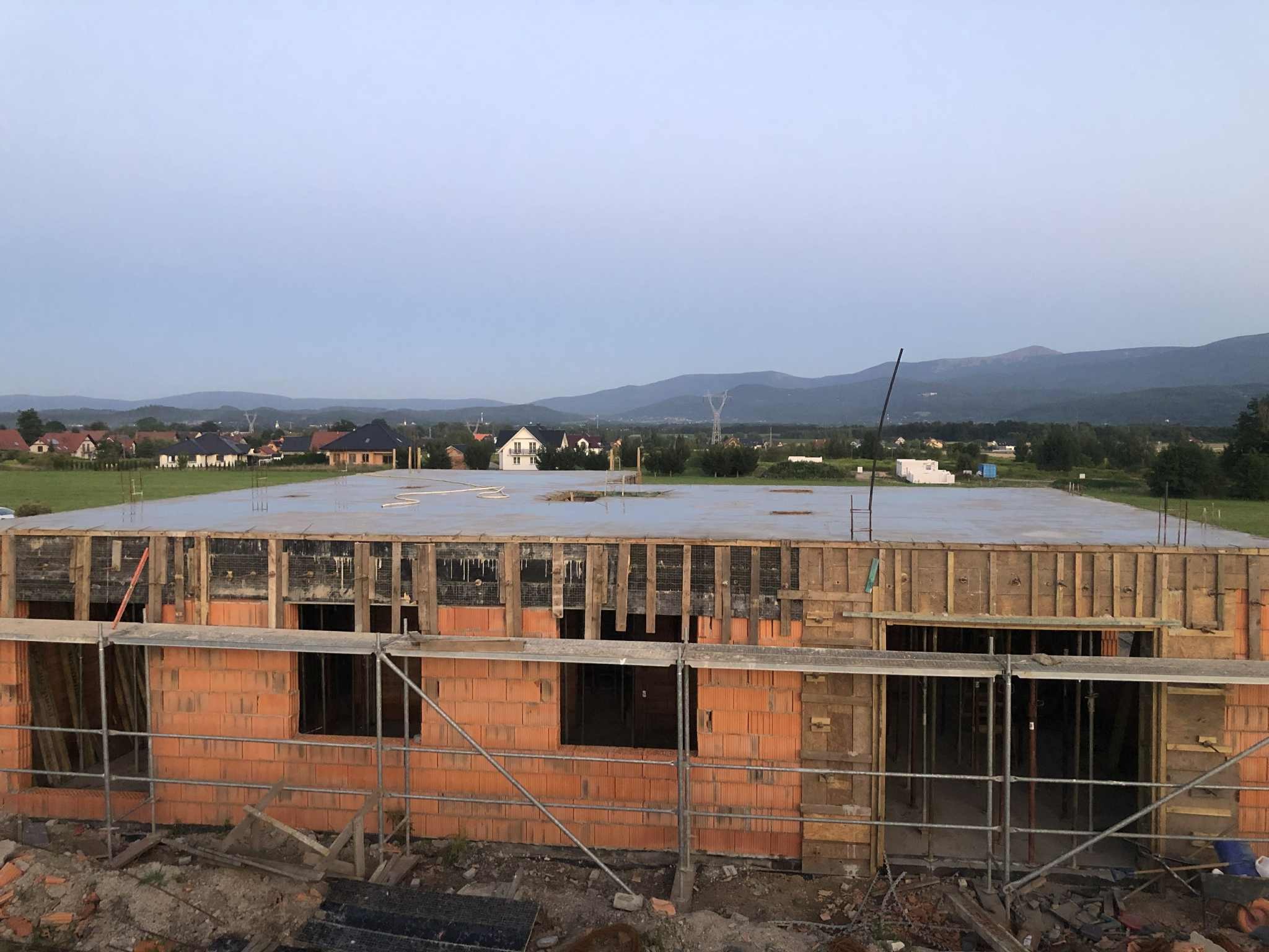 Budowa domu jednorodzinnego w trakcie realizacji: murowane ściany z pustaków, szalunek stropu, rusztowanie. W tle krajobraz z górami i zabudowaniami.