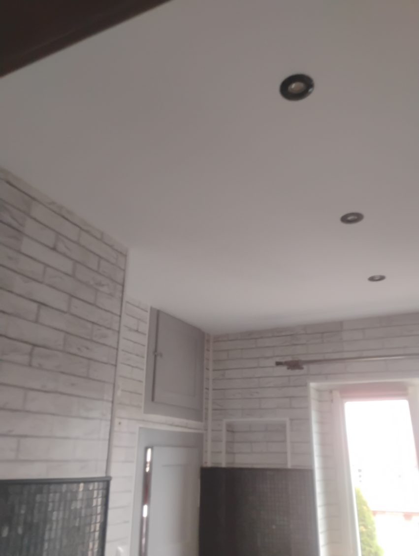 Wykończona łazienka z białymi płytkami imitującymi cegłę, oświetleniem punktowym i szarą szafką wnękową. Widok na okno z fragmentem zieleni na zewnątrz.