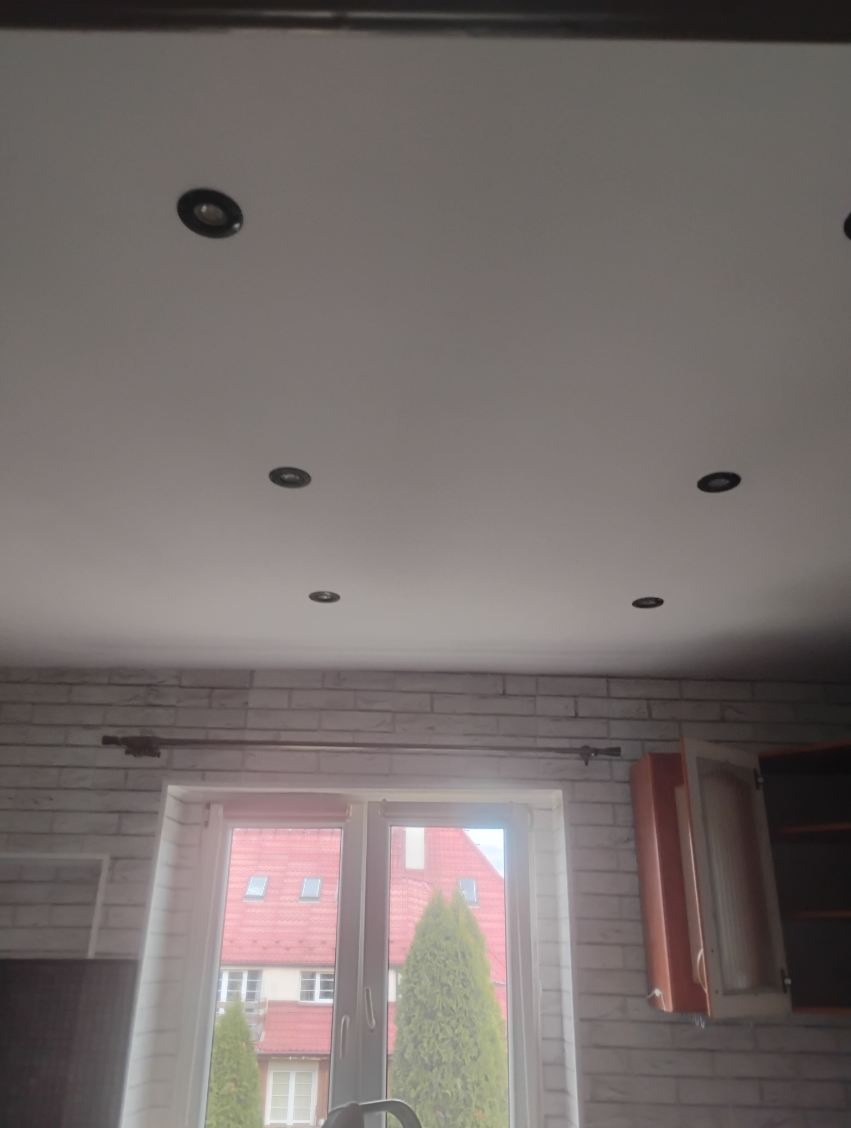 Biały sufit z sześcioma punktowymi lampami, ściana z białej cegły, okno z widokiem na budynek z czerwonym dachem i zielone drzewa. Częściowo widoczna szafka.