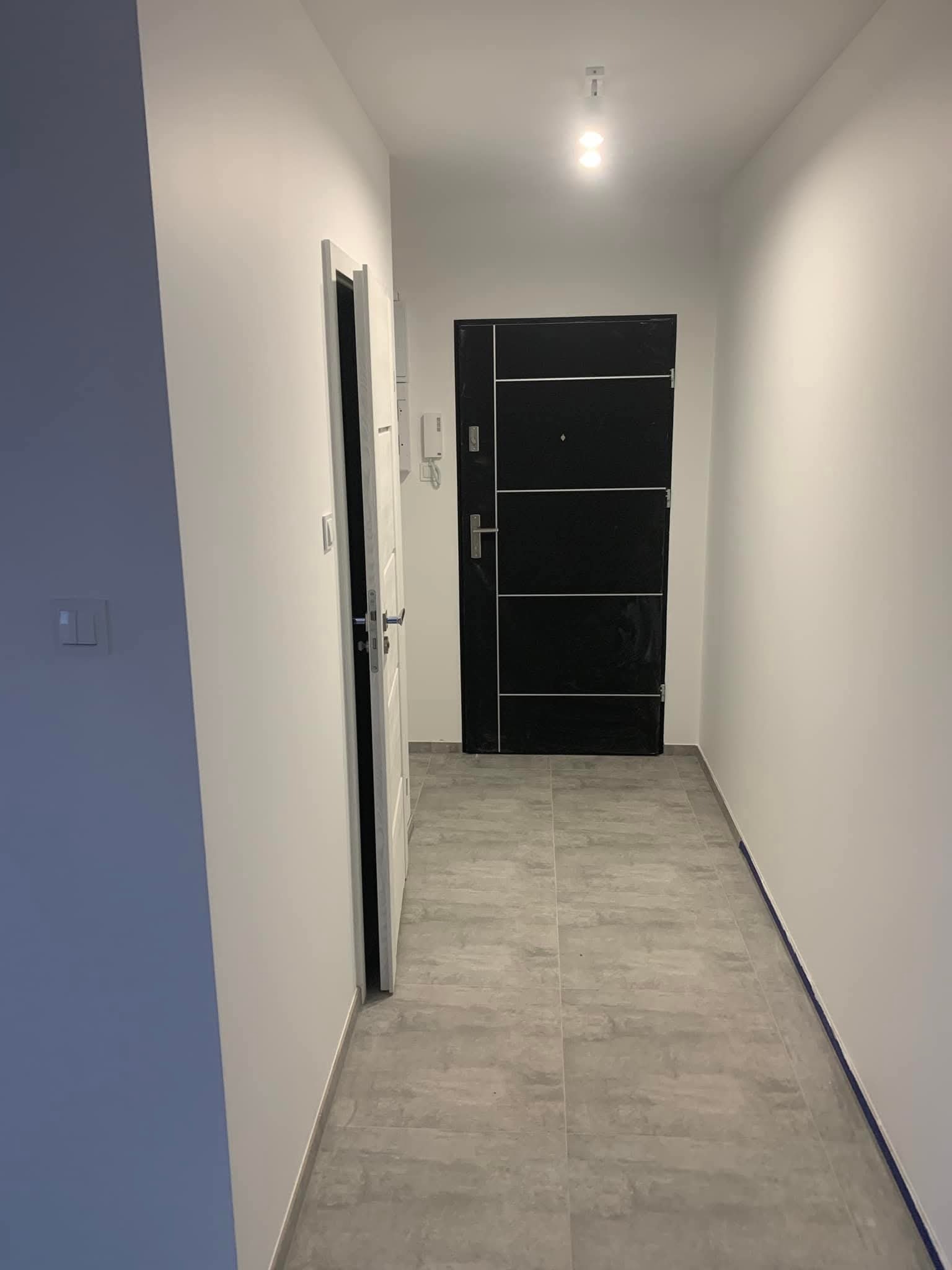 Korytarz po remoncie: szare płytki podłogowe, białe ściany, czarne drzwi wejściowe z poziomymi srebrnymi paskami, otwarte białe drzwi po lewej stronie.