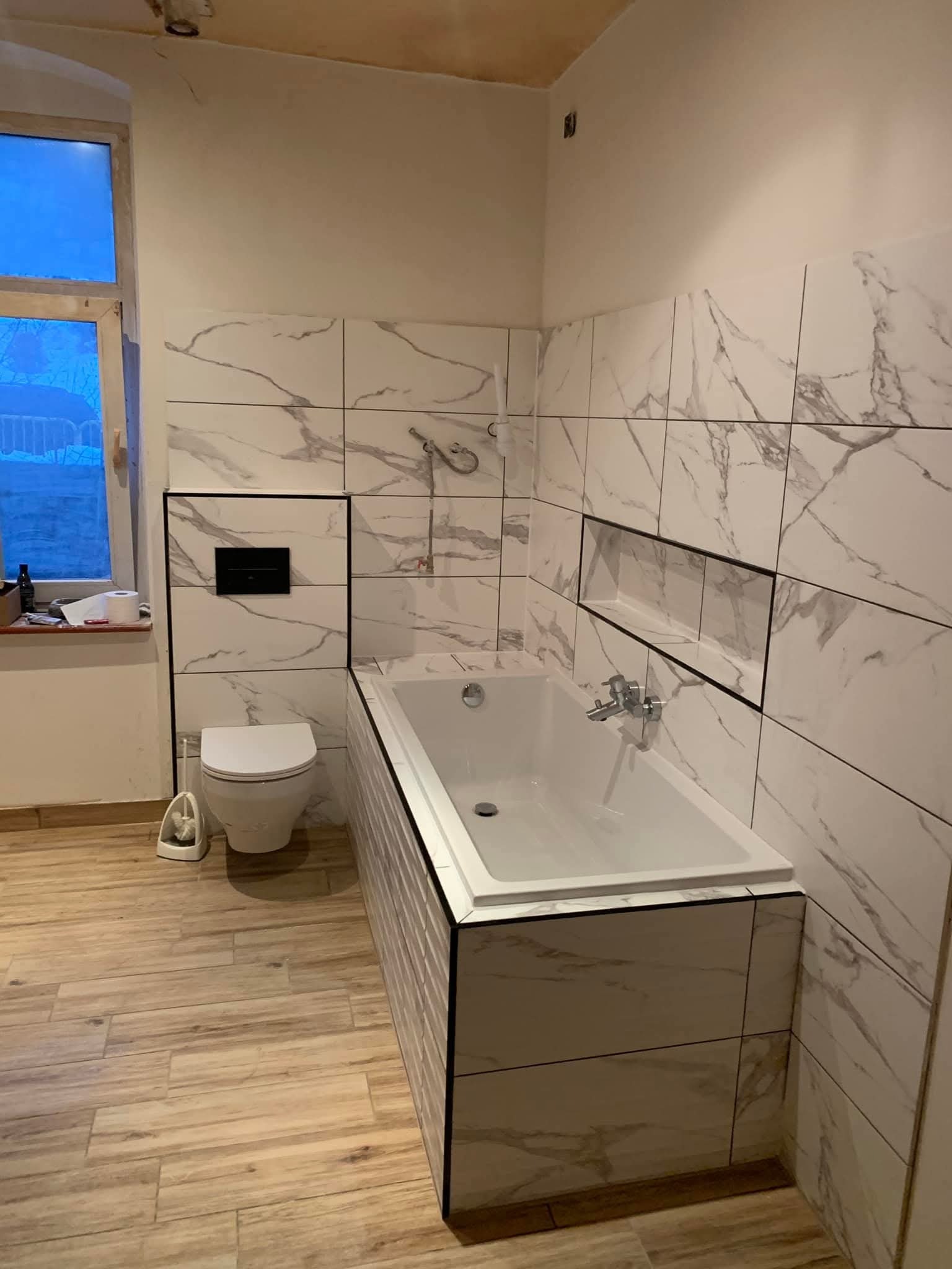 Wykończona łazienka z wanną obudowaną płytkami imitującymi marmur, toaletą podwieszaną i drewnopodobną podłogą. Widoczne okno z naturalnym światłem.