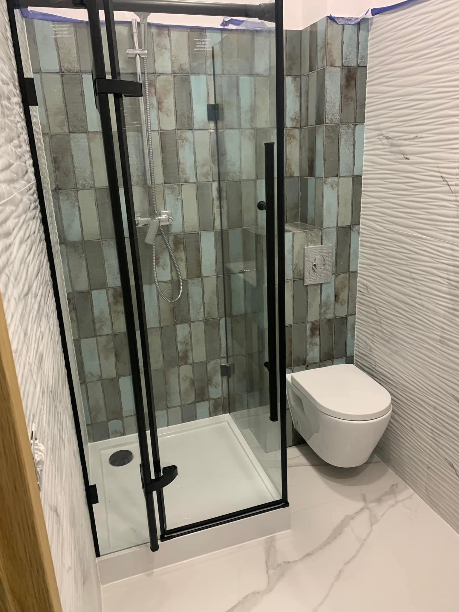 Nowoczesna łazienka z kabiną prysznicową typu walk-in z czarnymi profilami, płytkami w odcieniach szarości i błękitu oraz białą, wzorzystą ścianą i podwieszaną toaletą.