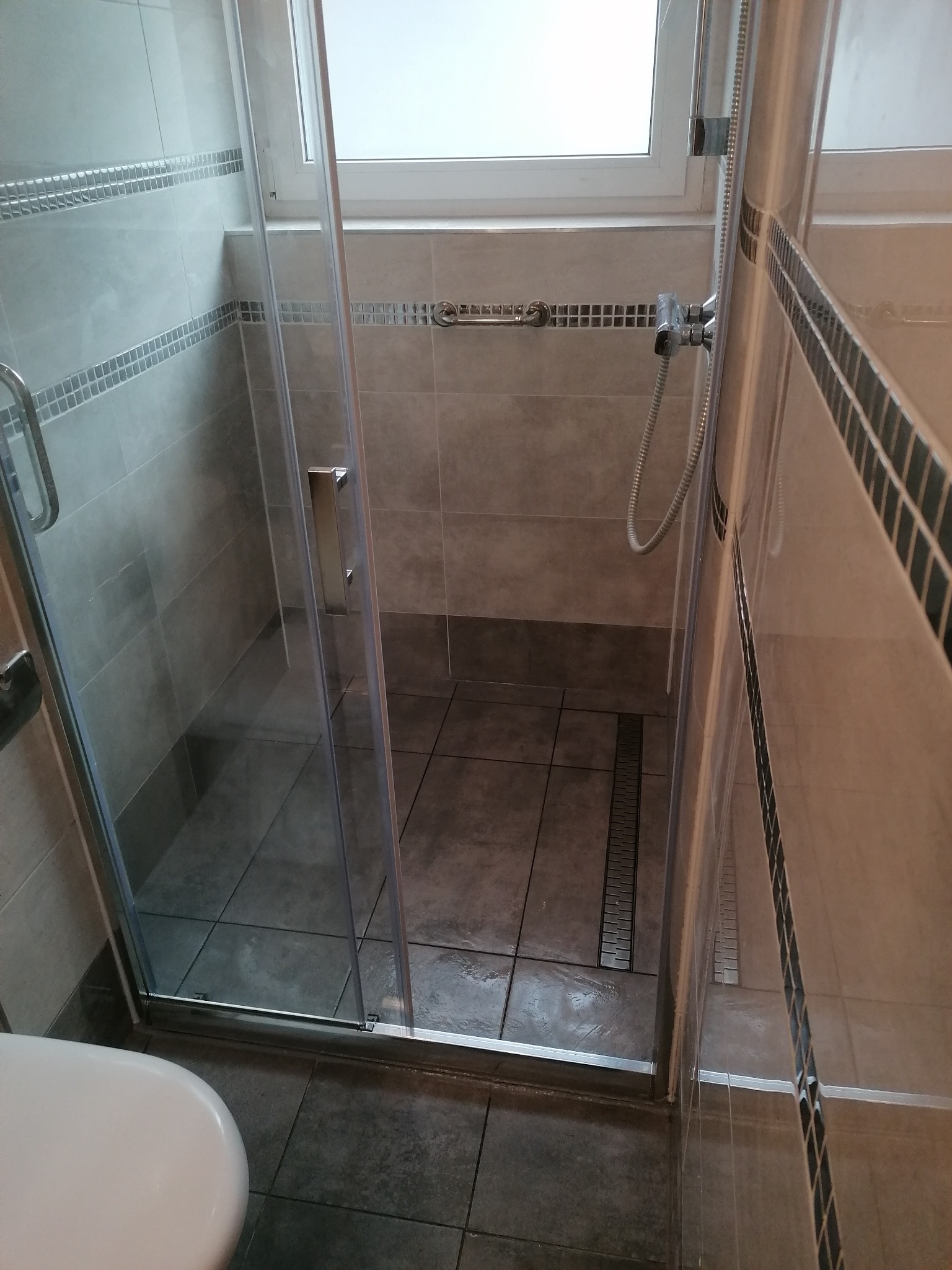 Nowoczesna kabina prysznicowa z odpływem liniowym, wykończona szarymi płytkami z dekoracyjnym pasem mozaiki. Widoczny fragment toalety.