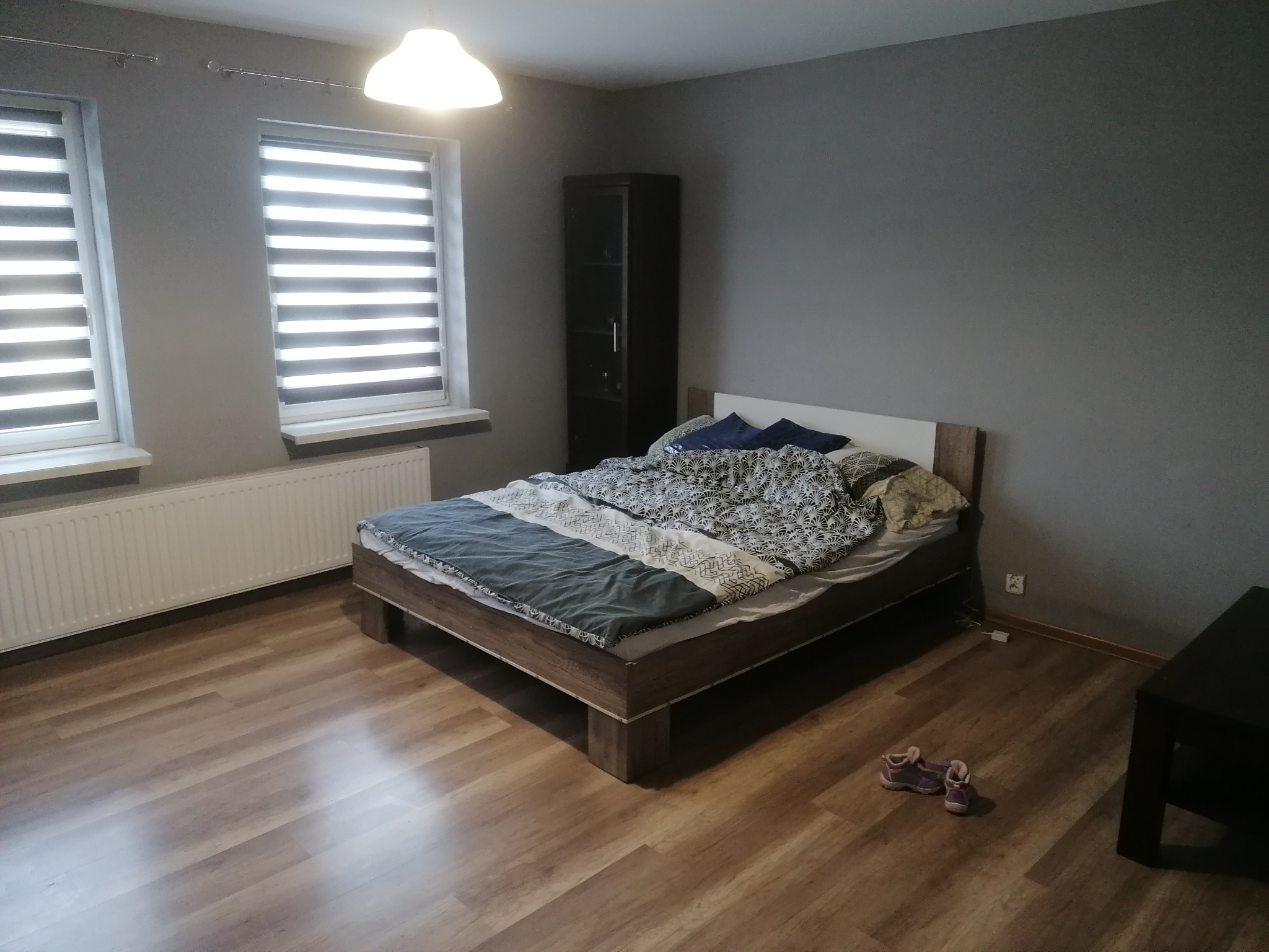 Pokój z drewnianą podłogą i łóżkiem. Dwa okna z żaluzjami dzień-noc wpuszczają światło. Ściany w odcieniach szarości. Minimalistyczny wystrój.