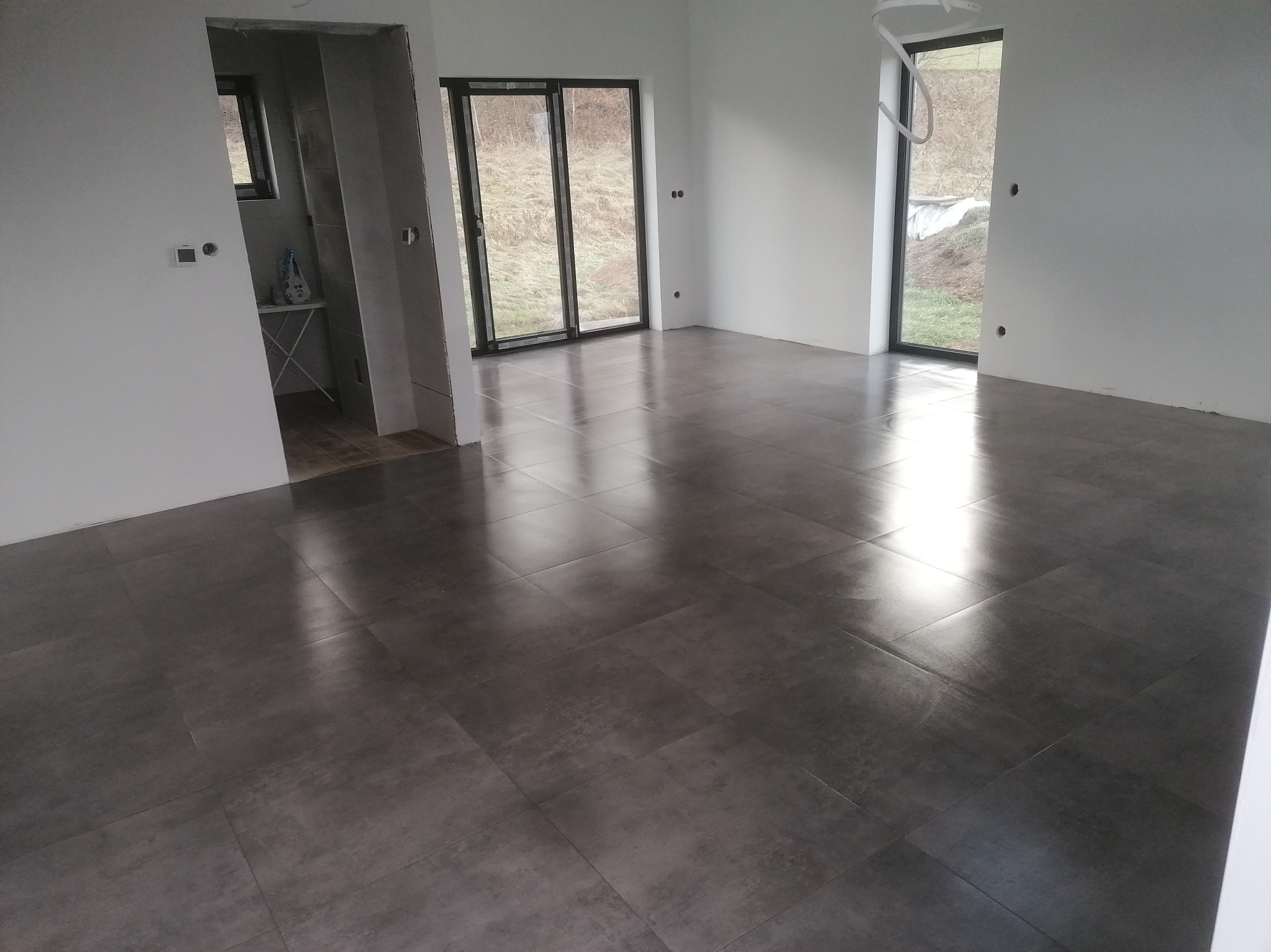 Nowoczesne, szare płytki podłogowe w przestronnym, jasnym wnętrzu z dużymi oknami. Widoczne przygotowania do wykończenia, surowe ściany i wystające kable.