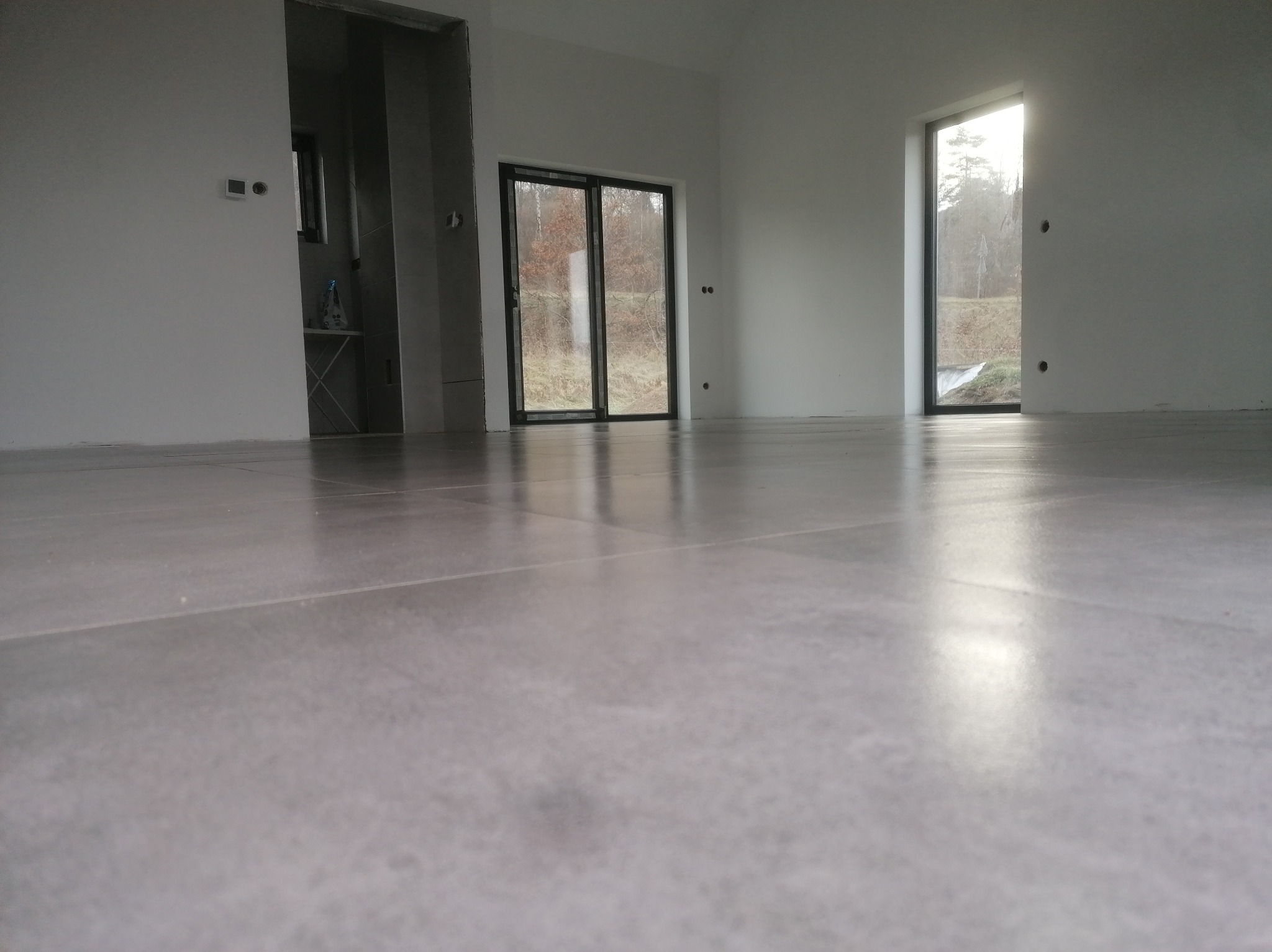 Jasnoszara, błyszcząca podłoga z płytek w nowym, minimalistycznym wnętrzu z dużymi oknami i drzwiami balkonowymi. Widoczny fragment łazienki w tle.