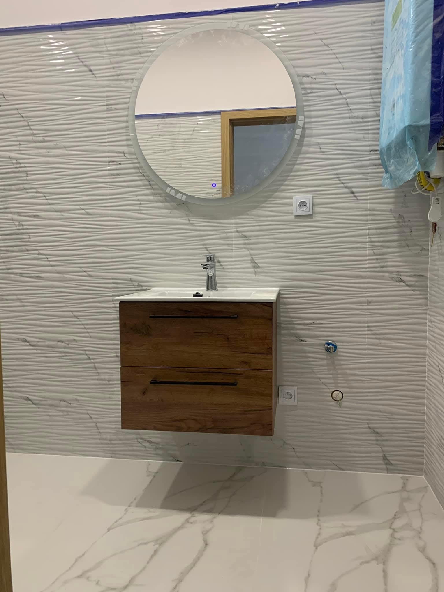 Nowoczesna łazienka z umywalką na szafce z drewnianym frontem, okrągłym lustrem z podświetleniem i płytkami 3D na ścianie. Widoczne gniazdka elektryczne i armatura.