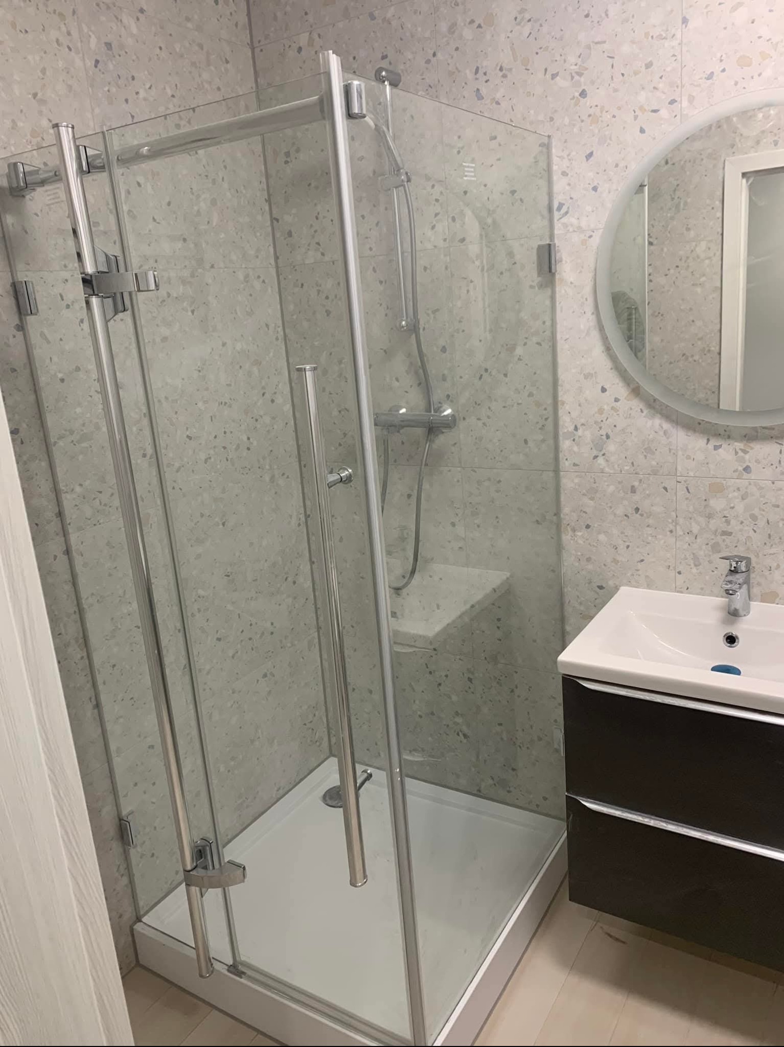 Nowoczesna kabina prysznicowa ze srebrnymi profilami i szklanymi ściankami, umywalka z szafką w ciemnym kolorze, okrągłe lustro i płytki w jasnych odcieniach.