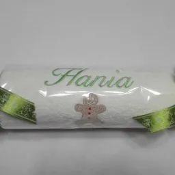 Biały ręcznik z zielonym haftem imienia 'Hania' i motywem piernikowego ludzika, zapakowany w folię i ozdobiony zielonymi wstążkami z motywami świątecznymi, przypominający kształtem cukierek.